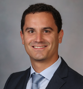 Portrait photo of a smiling man with short dark hair, wearing a dark grey suit jacket, a light grey shirt and a grey tie