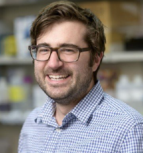 Portrait photo of a smiling man with wavy brown hair and eyeglasses wearing a blue checkered shirt with blurred shelves in the background