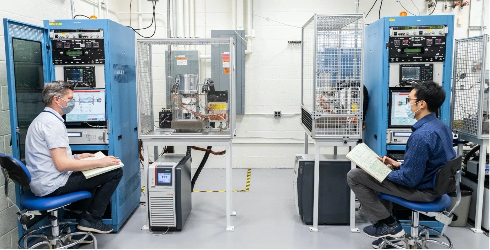 Photograph of two men wearing breathing masks and sitting in a lab with blue metal cases holding computer equipment.