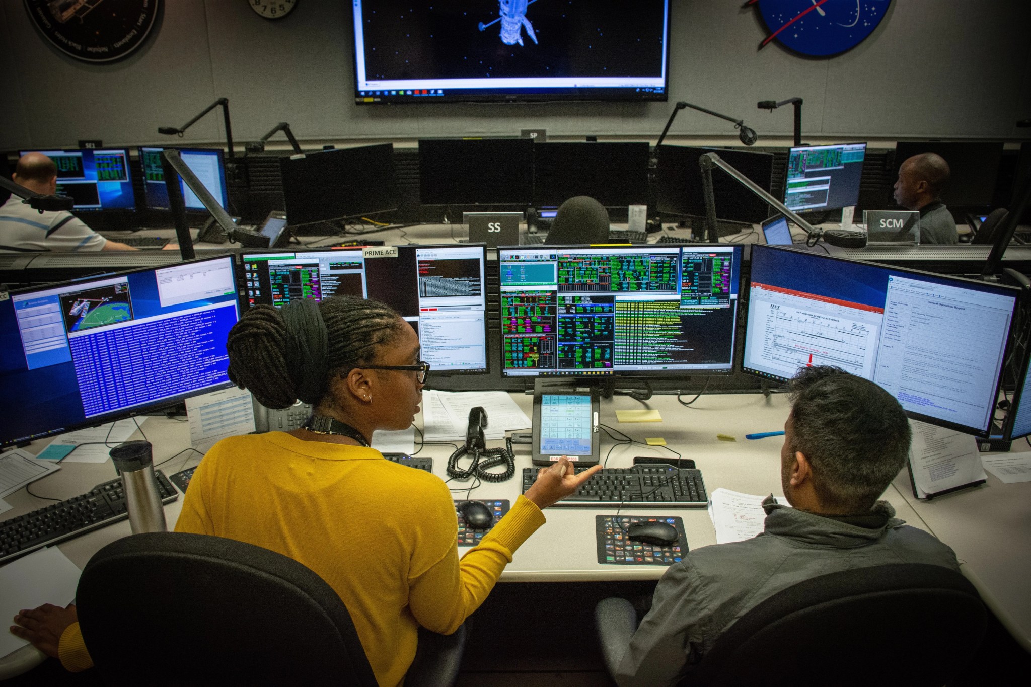 Hubble mission engineer Phanthom Donald, a Black woman with long black dreadlocks in a large bun on the back of her head, gestures and speaks to a fellow engineer sitting in front of several large computer monitors.