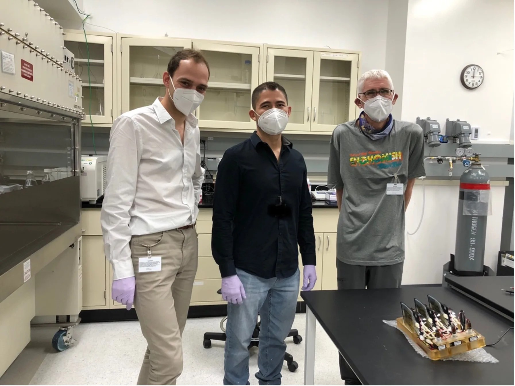 Three men wearing face masks and lavender colored latex gloves pose for a photo in a lab with a science component on the counter in front of them.