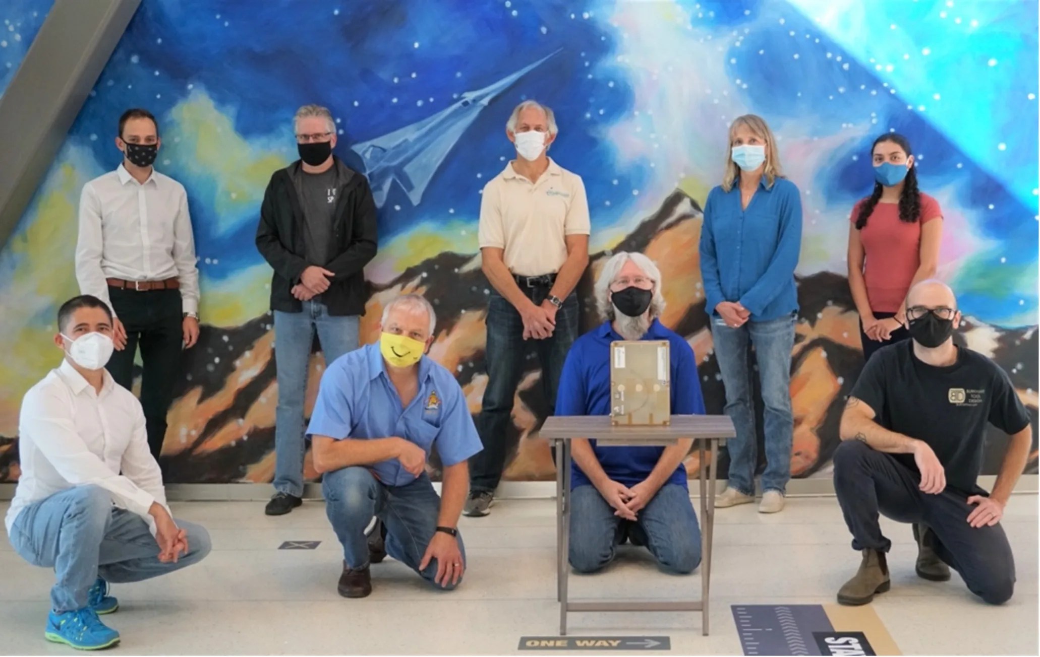 Group photo of 7 men and 2 women, 5 standing in the back and 4 kneeling in the front. The background is a painted mural of a spacecraft above rocky terrain with a blue background. In front of the group is a science component on a small table.