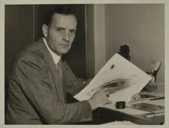 Edwin Hubble sitting at a desk holding a galaxy image.