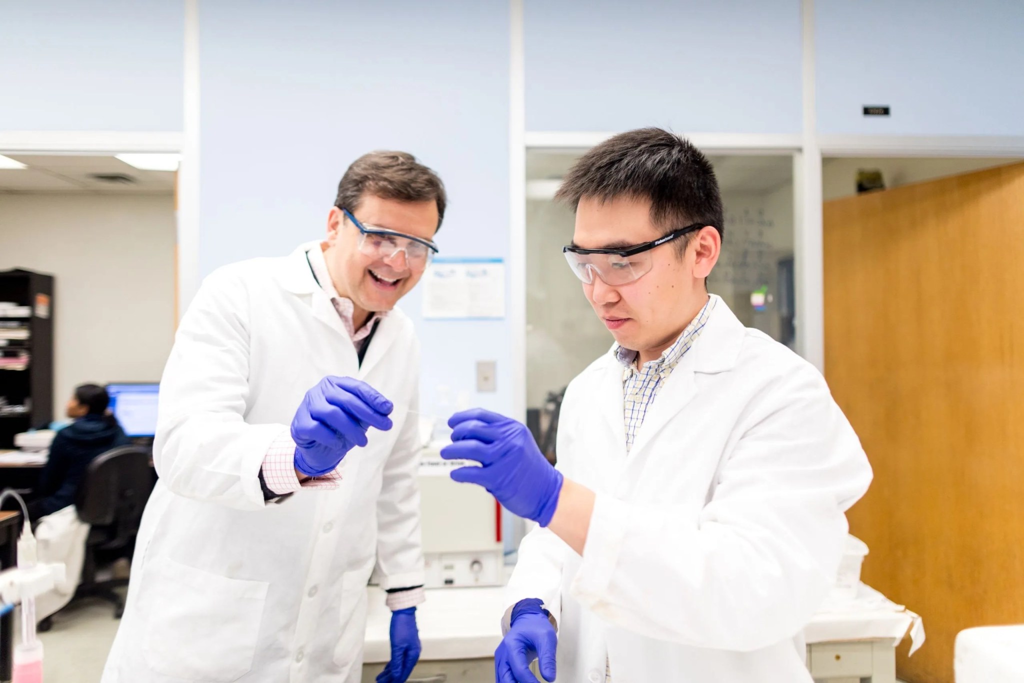2)Two men wearing lab coats, protective eyewear, and surgical gloves each hold one end of a long, slender tube.
