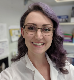 Portrait photo of a smiling young woman with purple died hair, eye glasses and wearing a white lab coat standing in an office
