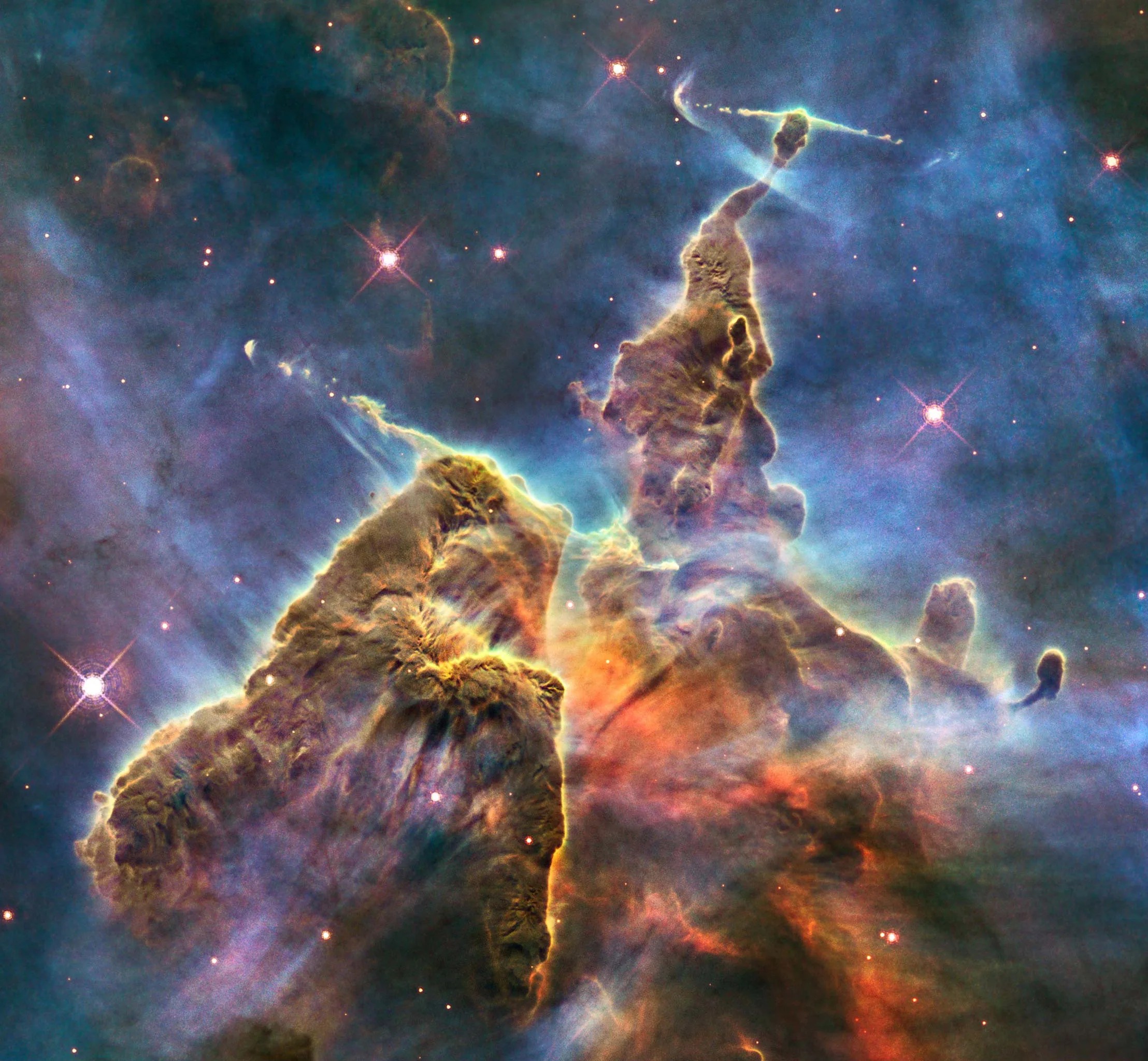 The Mystic Mountain is a chaotic, colorful pillar of gas and dust stretching up and narrowing toward the top of the image. The pillar is seen in mostly brown, yellow, and orange hues, jutting against a hazy blue and purple background dotted with a few pink stars.