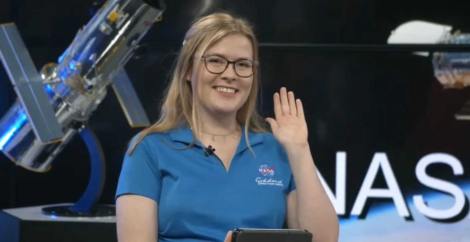 Elizabeth Tammi, a woman with glasses and long blonde hair, smiles and waves at the camera in this still image from a video.