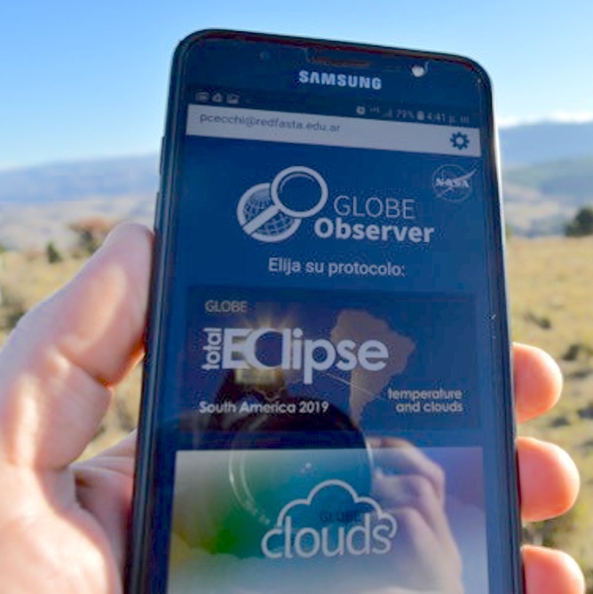Photo of a hand holding a mobile phone that is showing eclipse related citizen science graphics on the screen.