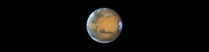 The planet Mars has an orange hue, splotched with dark brown areas of prominent features. A blue-white haze curves around the edges of the planet, particularly to the left in this image. A particularly bright spot of white clouds are visible to the right.