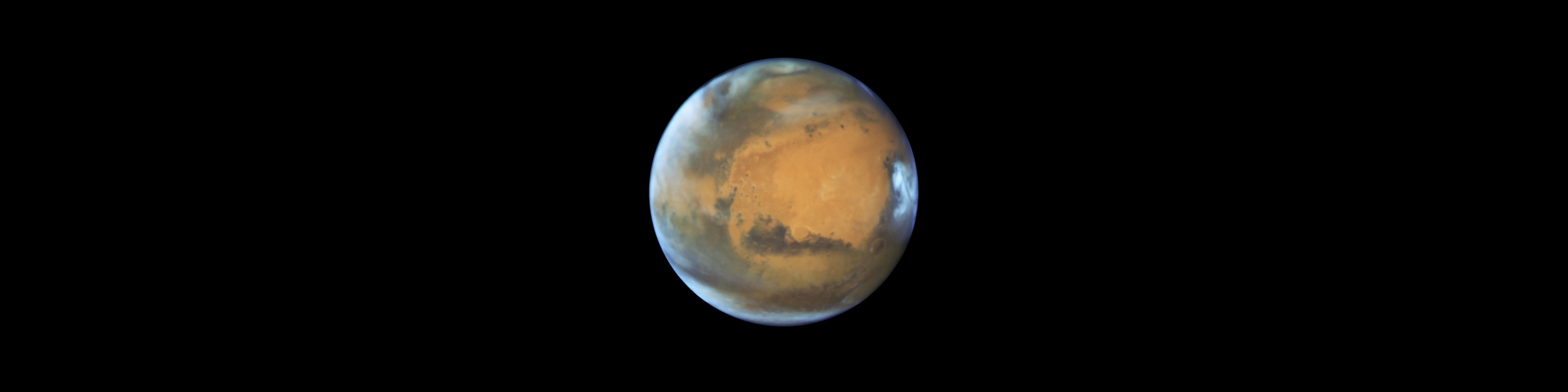 The planet Mars has an orange hue, splotched with dark brown areas of prominent features. A blue-white haze curves around the edges of the planet, particularly to the left in this image. A particularly bright spot of white clouds are visible to the right.