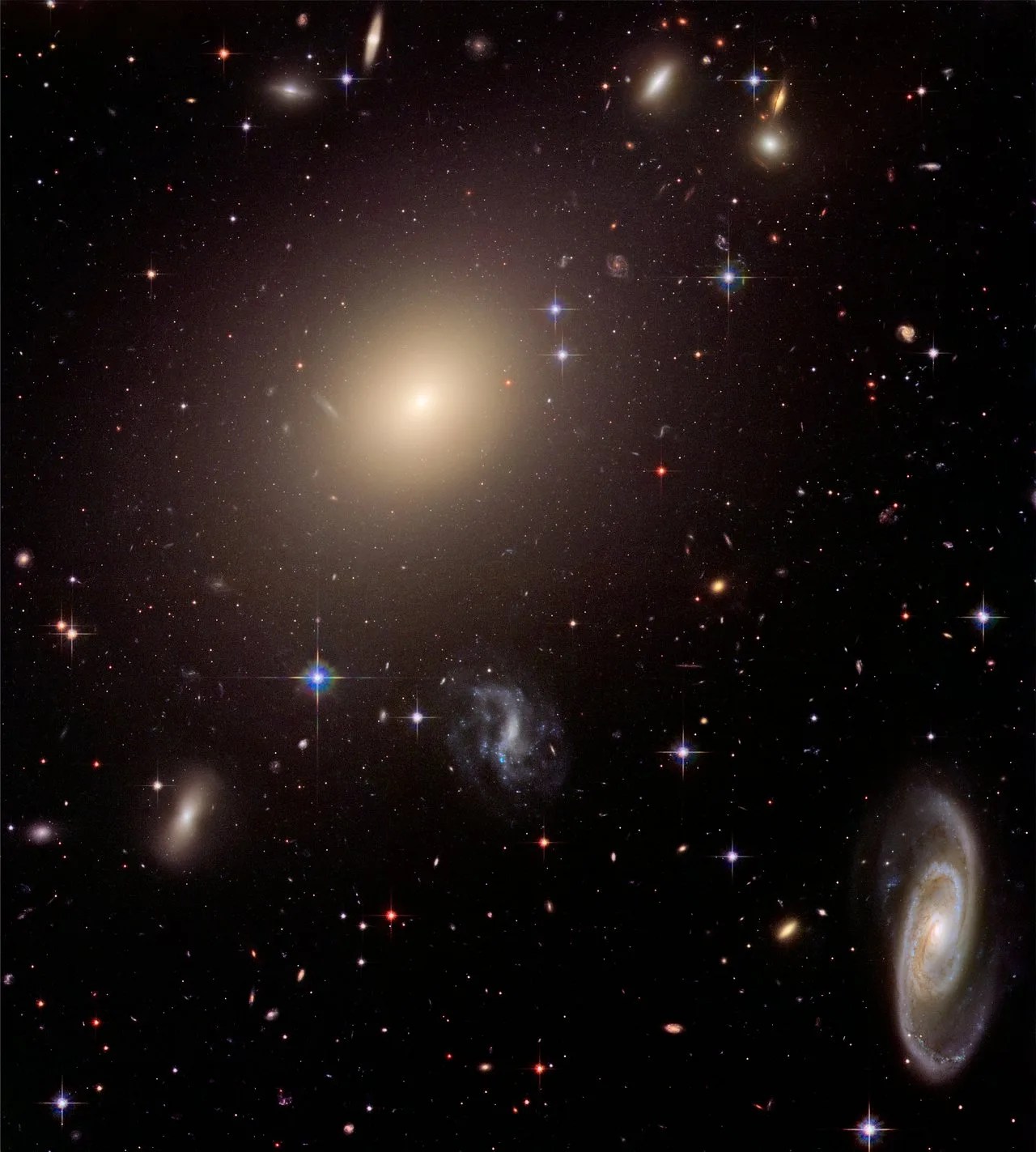 The field is filled with galaxies. One large elliptical galaxy is in the upper-left quadrant, just left of center. A large spiral galaxy is on its side near the right corner.