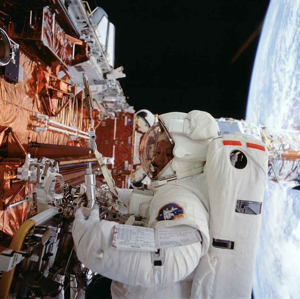 An astronaut in a space suit grips a tool while working on Hubble. The curve of Earth is visible in the background to the right.