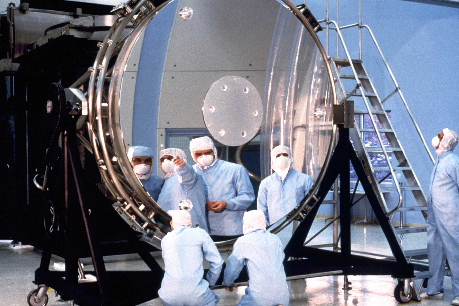 Technicians in blue suits, face masks, and head coverings look into Hubble's large primary mirror on a stand in a laboratory. They are reflected in the highly polished mirror.