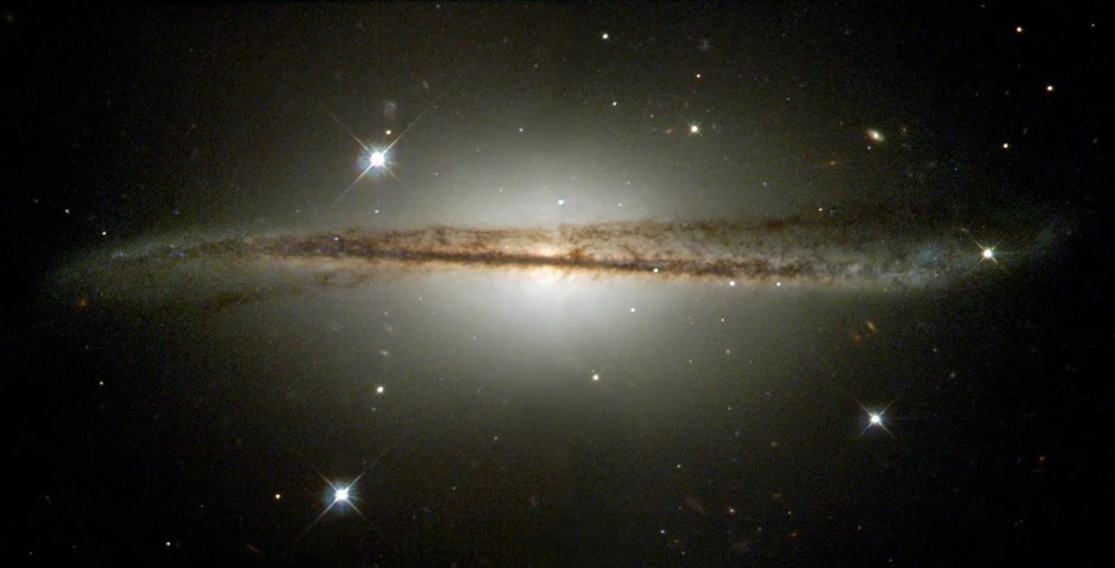 A bright ball of stars at image center. Along the equator of the ball is a warped disk of dust that appears like a dark, gentle wave across the ball of stars. Black background with a smattering of stars.