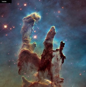 Three pillars of gas and dust rise from the bottom of the image. They are rusty-brown and black with golden highlights. A bright, hazy background of varying shades of blue and turquoise helps silhouette the darker pillars. Bright stars dot the background.