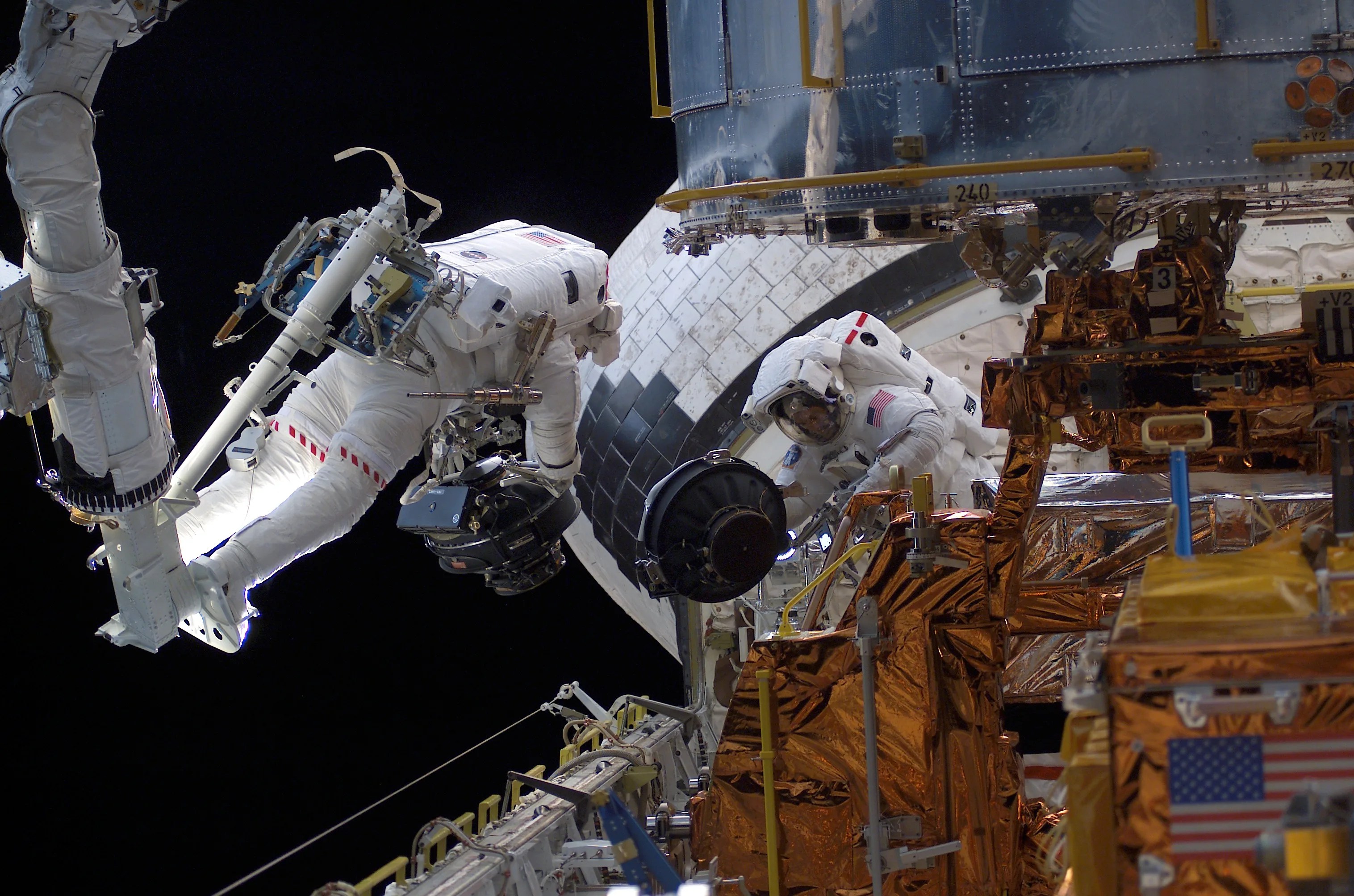 Two spacewalking astronauts, one standing attached to the space shuttle's robotic arm, work on the Hubble Space Telescope in the shuttle's cargo bay.