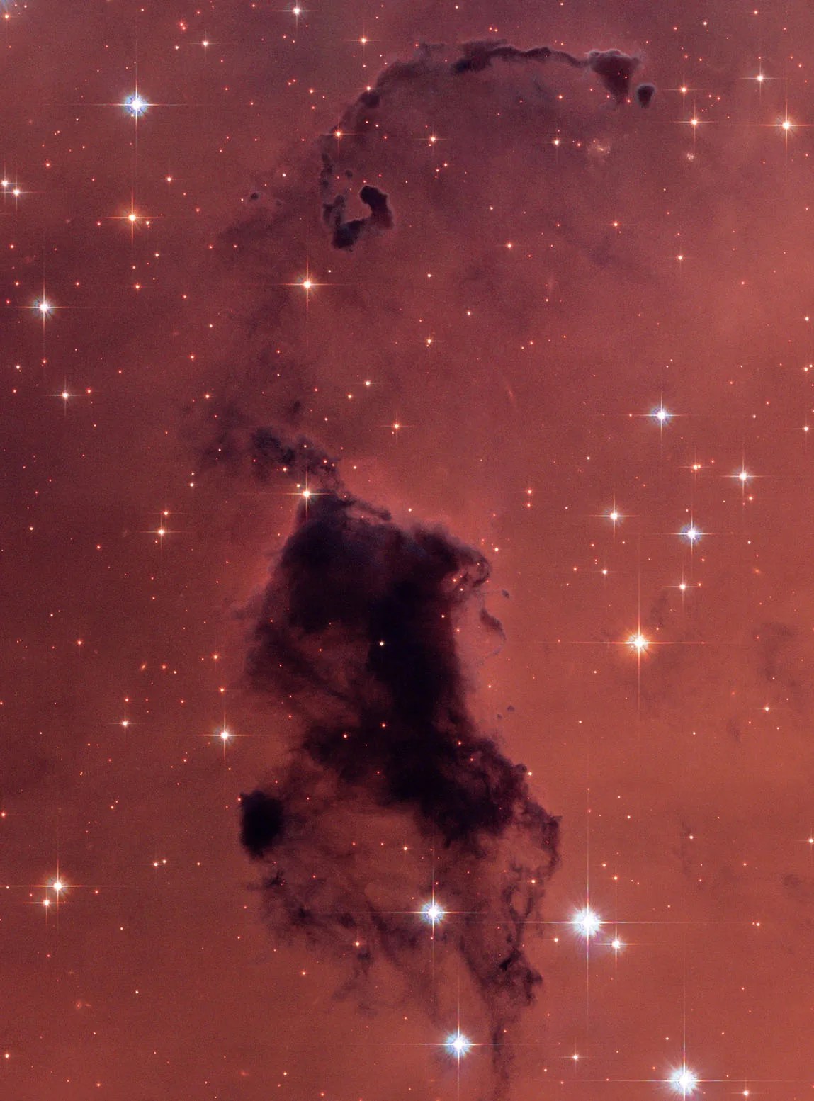 Glowing reddish gas is speckled with bright-white stars. In the foreground is a dark, vertically extended cloud of gas and dust that blocks the light of objects behind it. The largest part of the dark cloud (Bok Globule) extends from image center to bottom. Tenuous clouds extend vertically above the main globule toward the image top. 