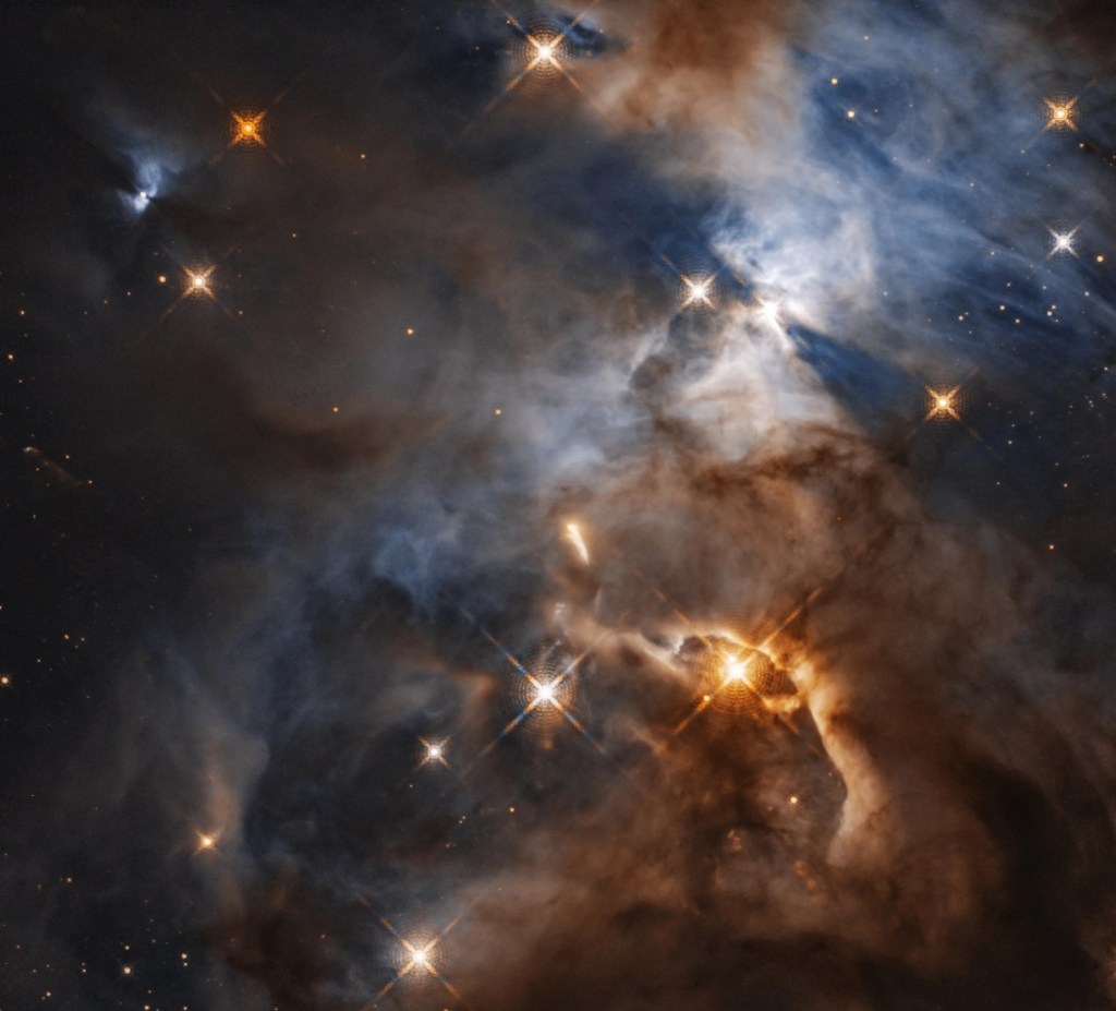 Dark clouds fill the scene. They are outlined by bright rusty-orange, and blue-white clouds of gas. Stars dot the scene.