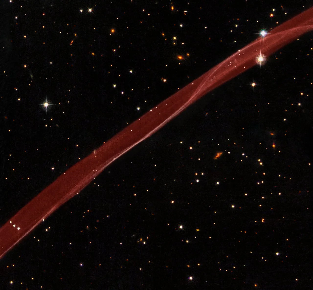 A black background dotted with stars is bisected from the lower-left corner to the upper-right corner by a slightly twisted strand of red gas and dust.