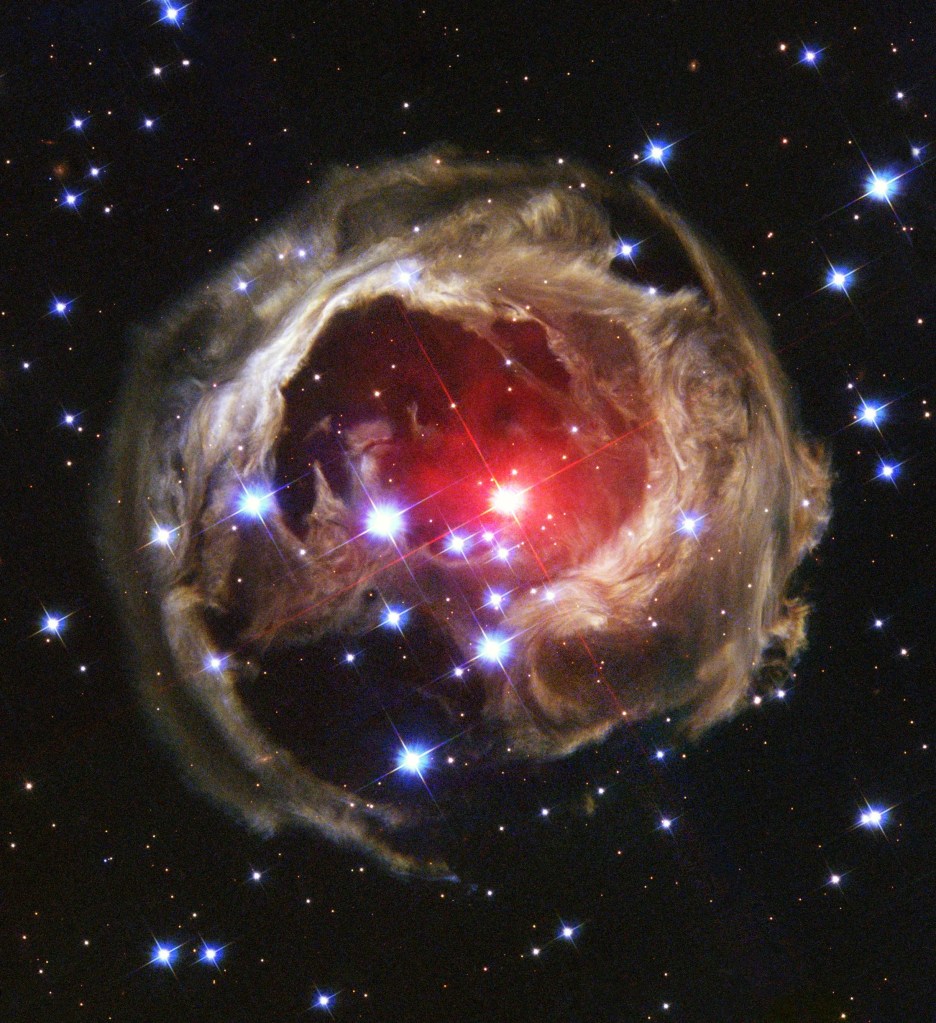 Hubble view of an expanding halo of light around star V838 Monocerotis. Center of the ball-like cloud holds a bright star surrounded by red gas. The outer region of the u0022ballu0022 is a tan color dotted with stars. Black background dotted with stars.