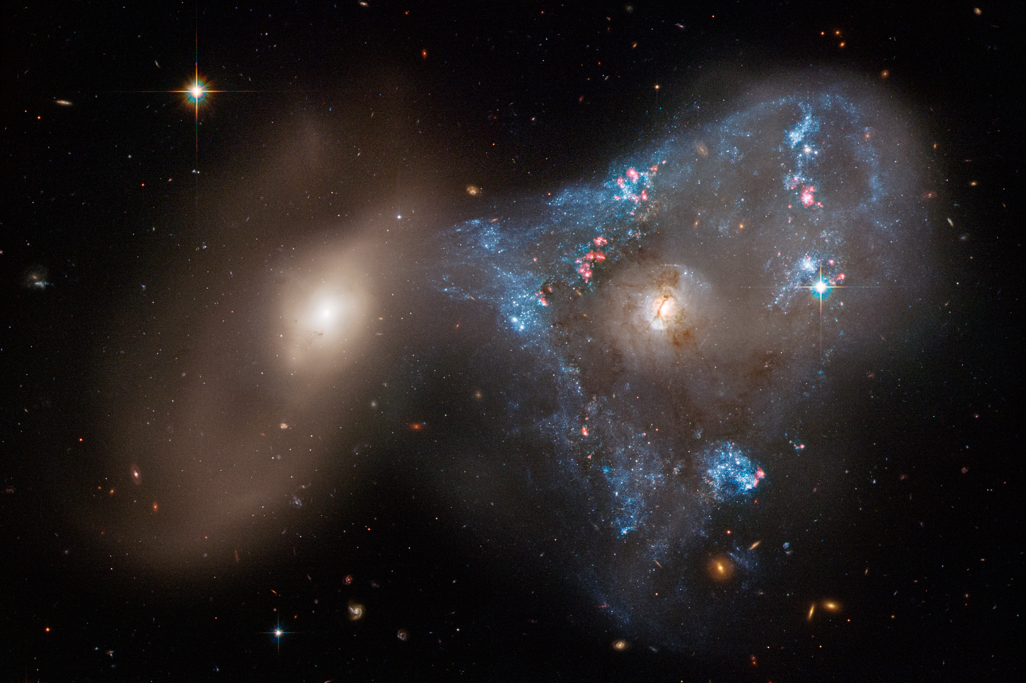 Two galaxies one right one on the left. right galaxy has a blue-white triangular star forming region surrounding it.