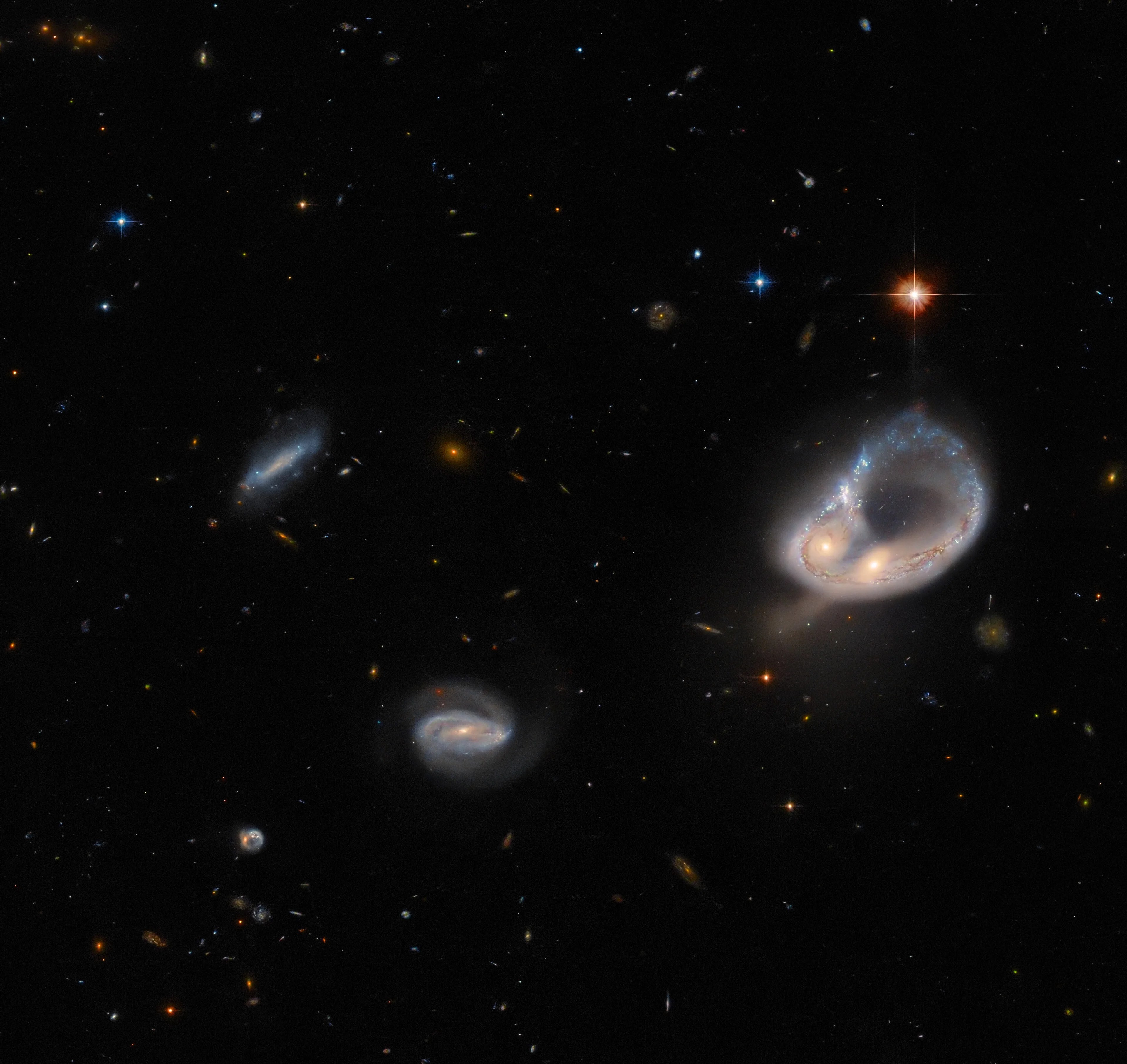 2 galaxies right of center form a narrow, blue ring. the cores of the 2 galaxies form a bulge on the ring’s side. bright, orange star lies above the ring. 2 smaller spiral galaxies left of center. black background speckled with small stars and galaxies.