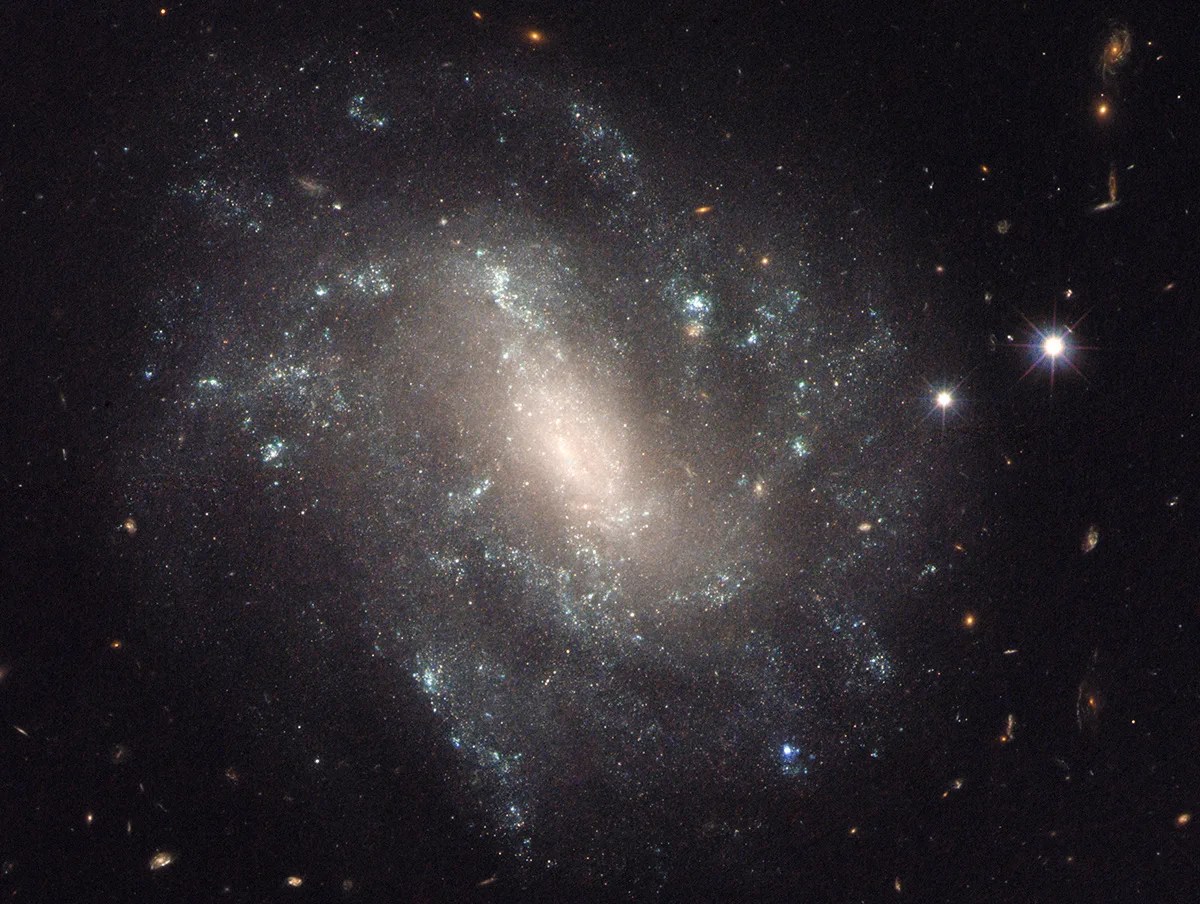 A sparkling galaxy with a bright, glowing core and patchy spiral arms dotted with pale blue star formation.