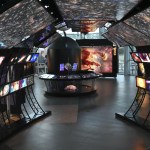 View of from within the entry tunnel of the Hubble exhibit. Illuminated Hubble images of galaxies and stars overhead, individual objects along the walls. Hubble Space Telescope model at the end of of the tunnel. Large illuminated Eagle Nebula image beyond.