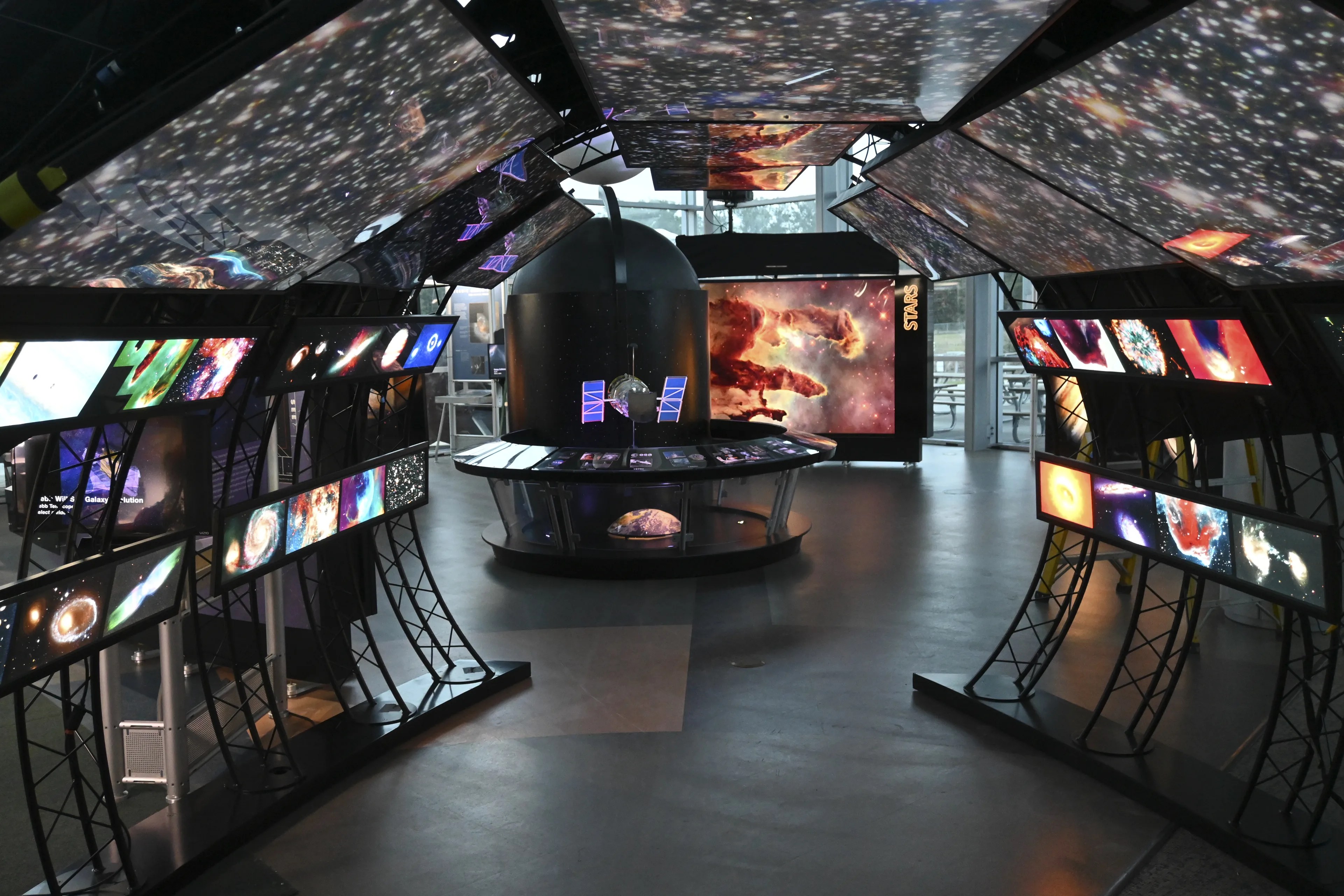 View of from within the entry tunnel of the Hubble exhibit. Illuminated Hubble images of galaxies and stars overhead, individual objects along the walls. Hubble Space Telescope model at the end of of the tunnel. Large illuminated Eagle Nebula image beyond.