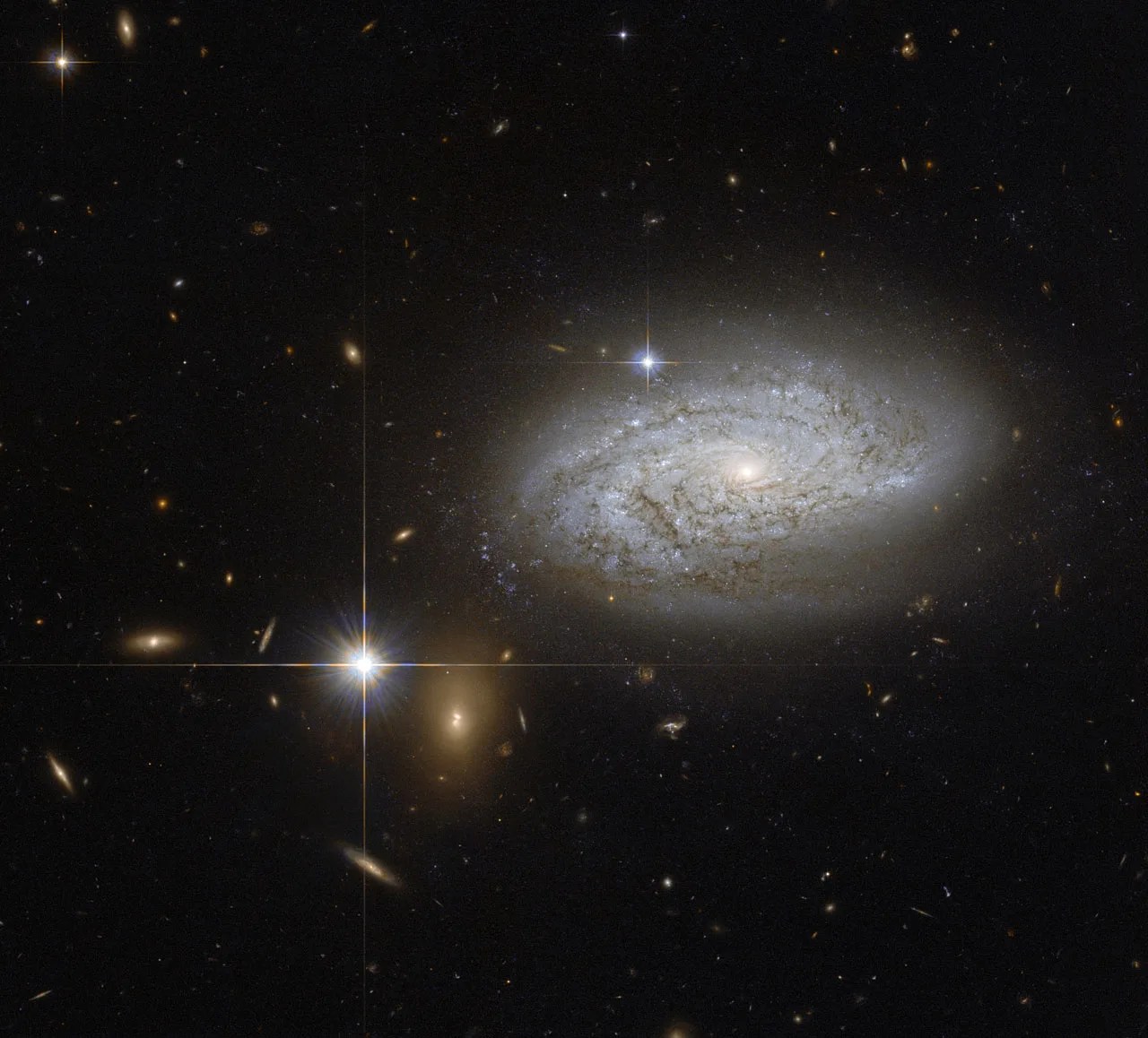 A spiral galaxy appears as a swirl of dust and light on a field of stars, with more galaxies in the background and a bright foreground star visible nearby.