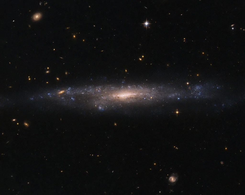 A galaxy is seen almost edge-on, with a bright orange-yellow core surrounded by patchy violet regions of star formation. The rest of the image is dotted with distant stars and galaxies against black space.