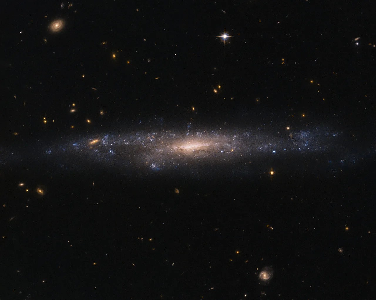 A galaxy is seen almost edge-on, with a bright orange-yellow core surrounded by patchy violet regions of star formation. The rest of the image is dotted with distant stars and galaxies against black space.