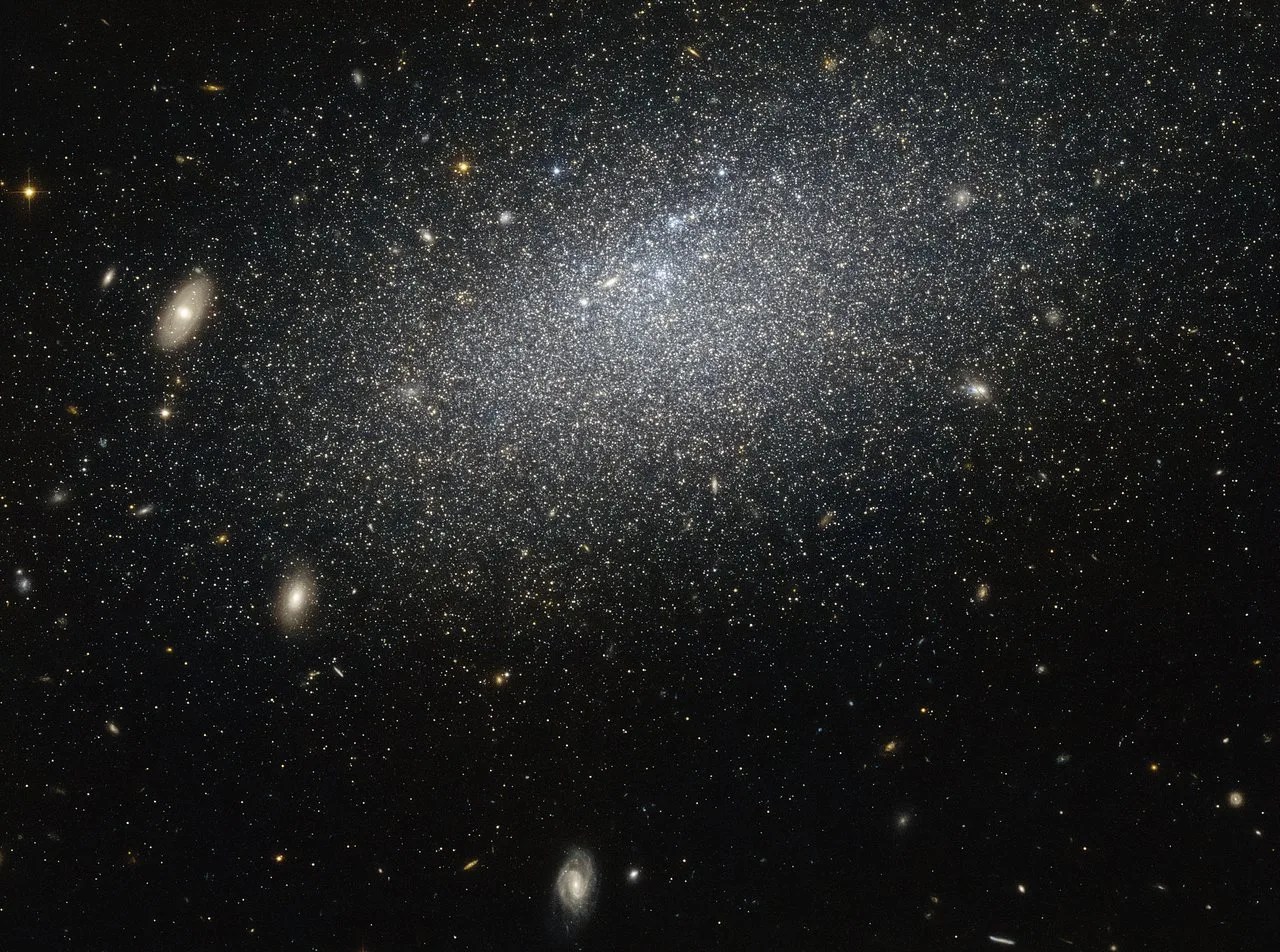 A thick blanket of stars spreads across the upper part of the image against the black background of space. The stars are thicker in the center of the grouping and more scattered in the outer regions. Smaller, more distant galaxies, including several obvious spirals, dot the background of the image.