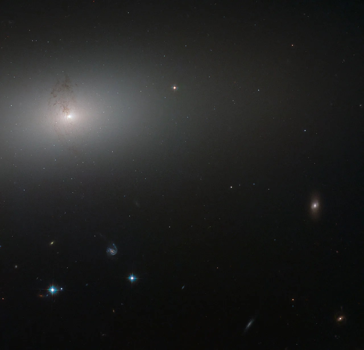 A distant elliptical galaxy is a soft white glow around a bright white core. The glow slowly fades out into the darkness of space, where a scattering of other smaller and more distant galaxies are visible. A swirl of dark dust lanes is visible agains the ellipitcal's glow.