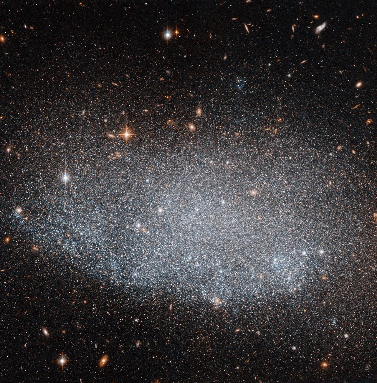 A large, irregular oval of stars fills the image. Some bright foreground stars and many smaller, more-distant galaxies are visible in the background.