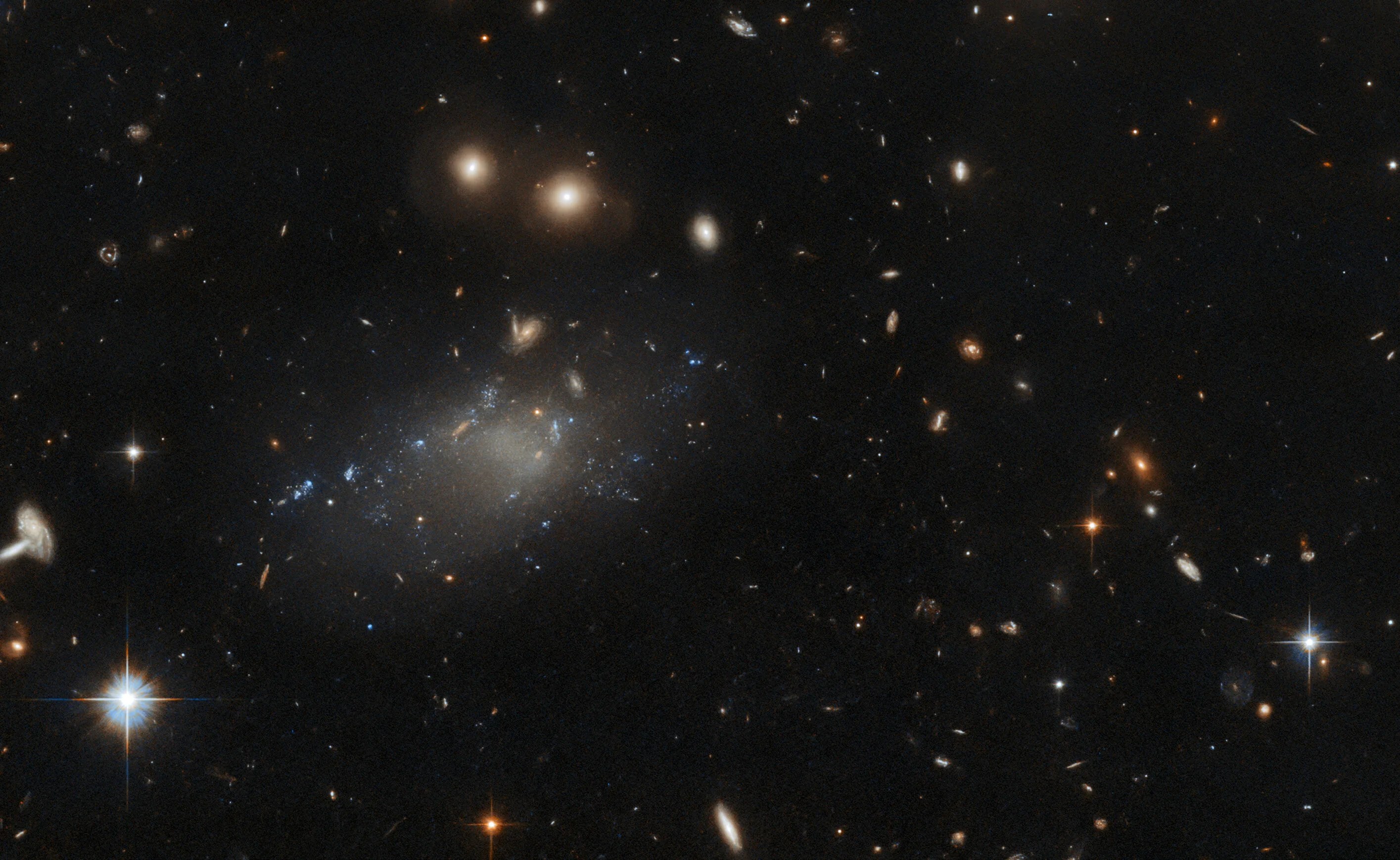 Cloud-like galaxy just left of center with many other galaxies filling the scene. bright foreground stars at lower left and lower right