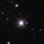 Black background peppered with blue-white stars and distant rusty-orange-red galaxies. center of image: bright star that holds a gaseous, blue, elongated, amorphous mass peppered with white stars just to the right of the star.