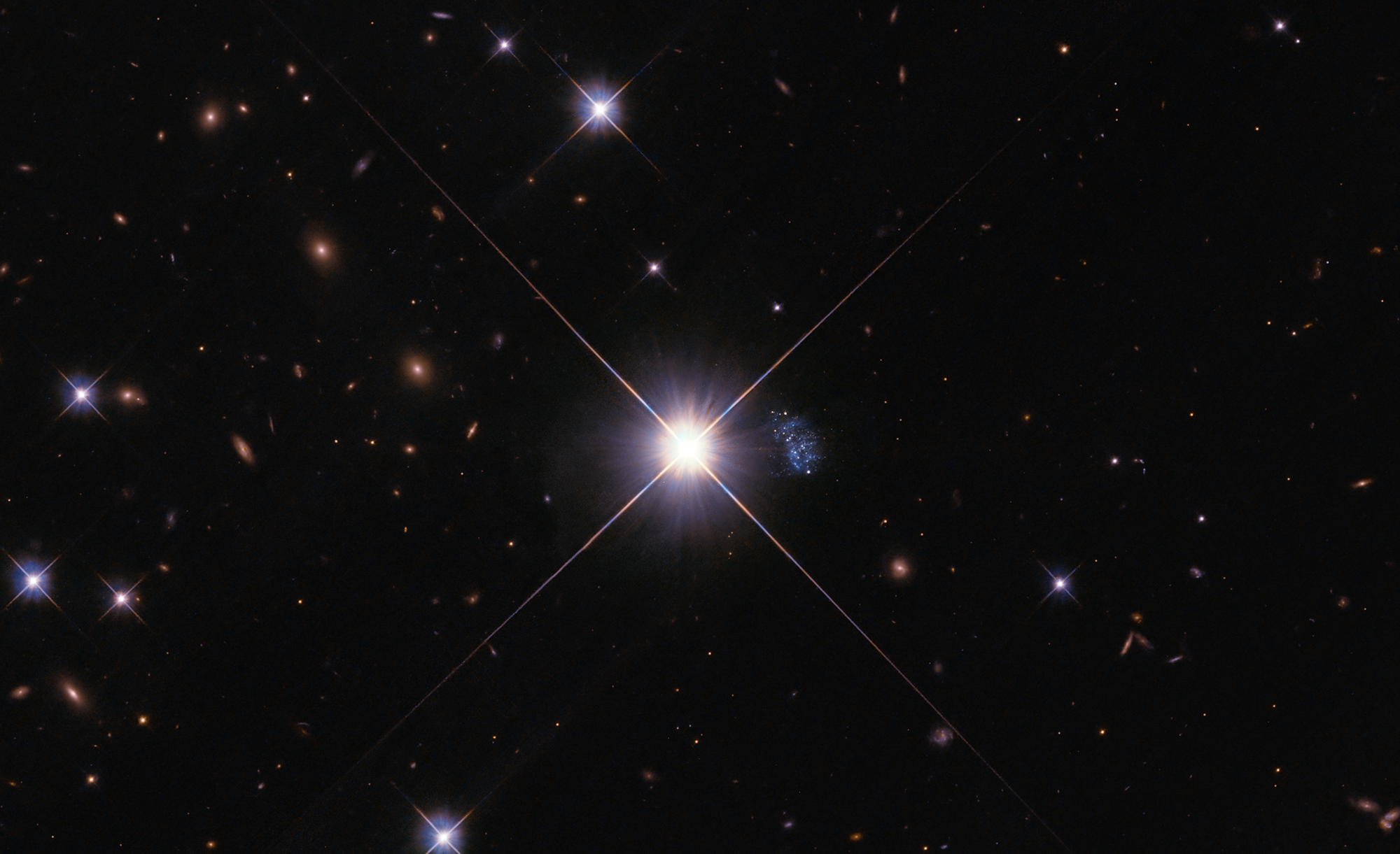 Black background peppered with blue-white stars and distant rusty-orange-red galaxies. center of image: bright star that holds a gaseous, blue, elongated, amorphous mass peppered with white stars just to the right of the star.