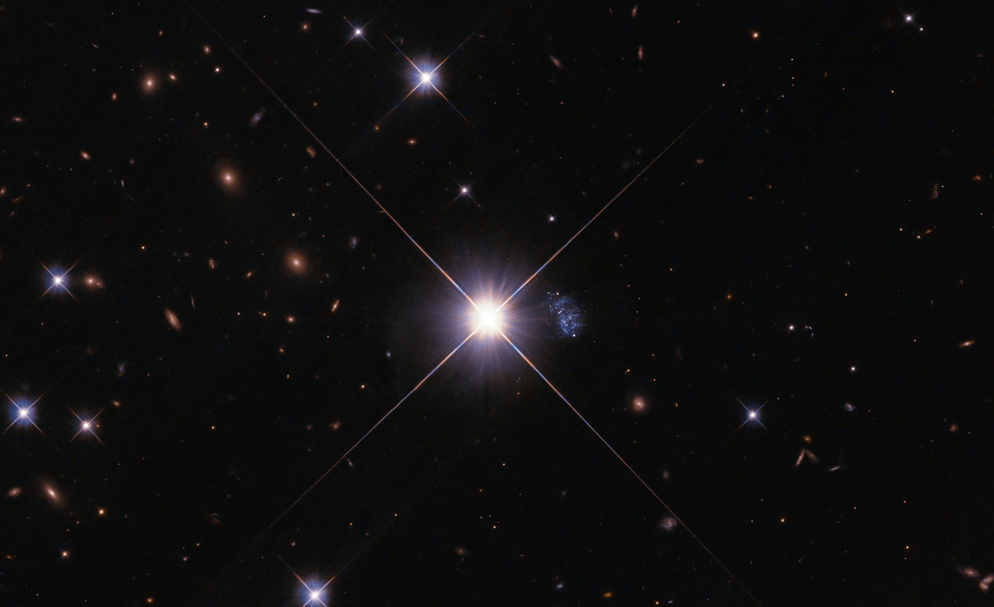 Black background peppered with blue-white stars and distant rusty-orange-red galaxies. center of image: bright star that holds a gaseous, blue, elongated, amorphous mass peppered with white stars just to the right of the star.
