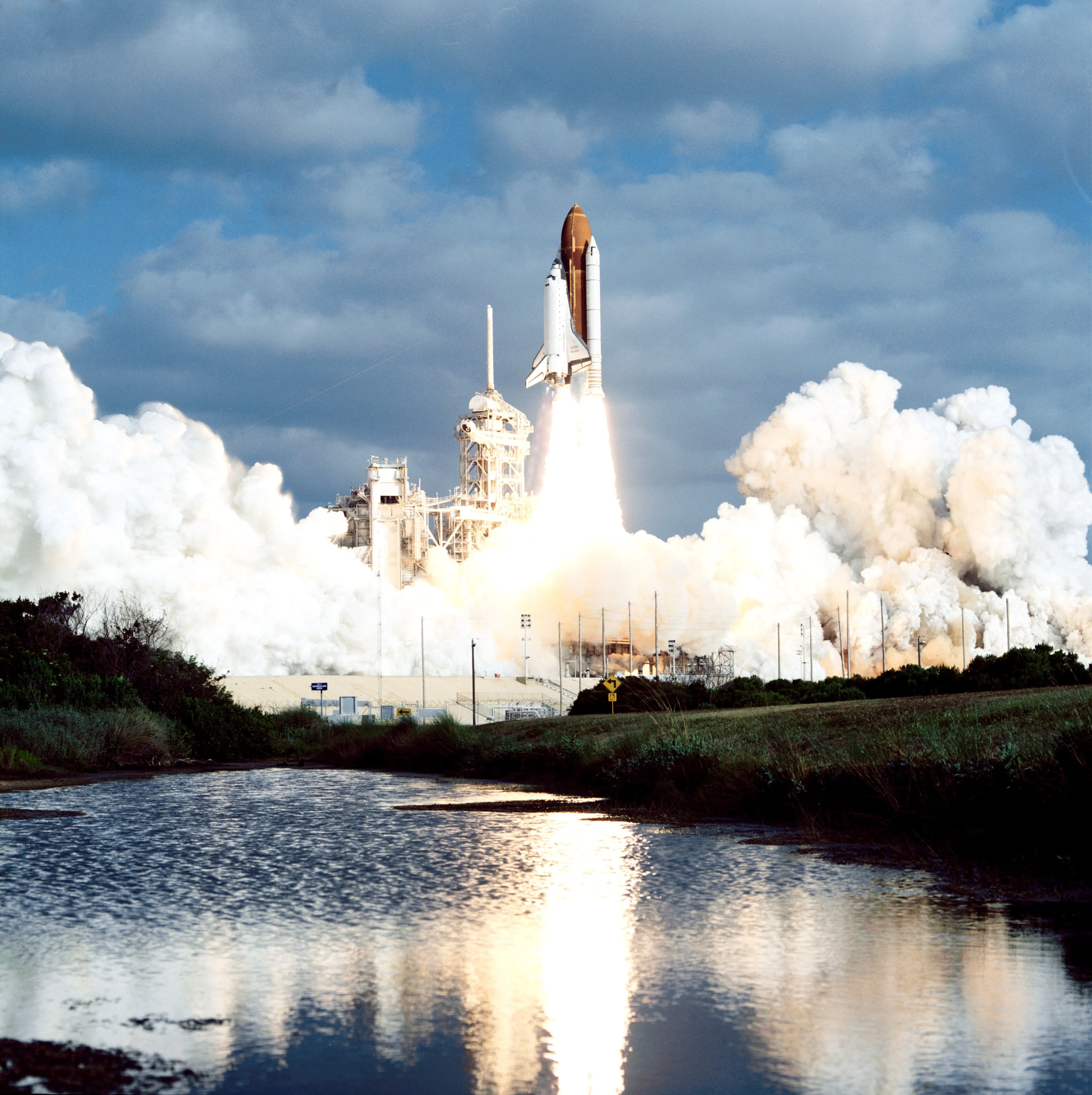 Clouds of steam and smoke billow as the Space Shuttle Discovery launches into blue skies, with Hubble aboard. the fire of the engines is reflected in a body of water in the foreground.