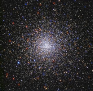Image center holds a dense sphere of stars in colors of red, orange, yellow, white, and blue. The stars are densely packed at image center and taper out toward the image's edges. They are on a black background.