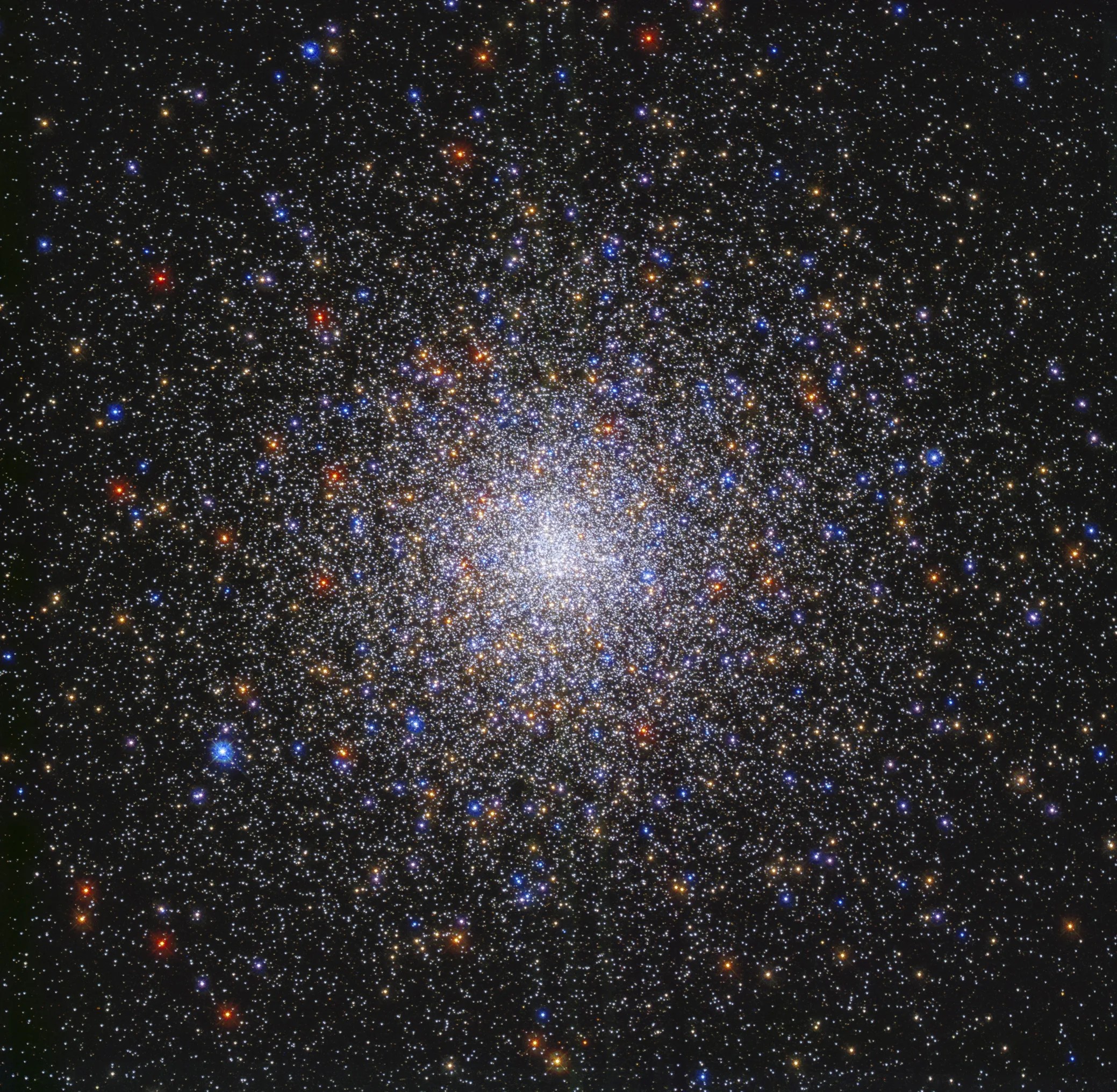 Image center holds a dense sphere of stars in colors of red, orange, yellow, white, and blue. The stars are densely packed at image center and taper out toward the image's edges. They are on a black background.
