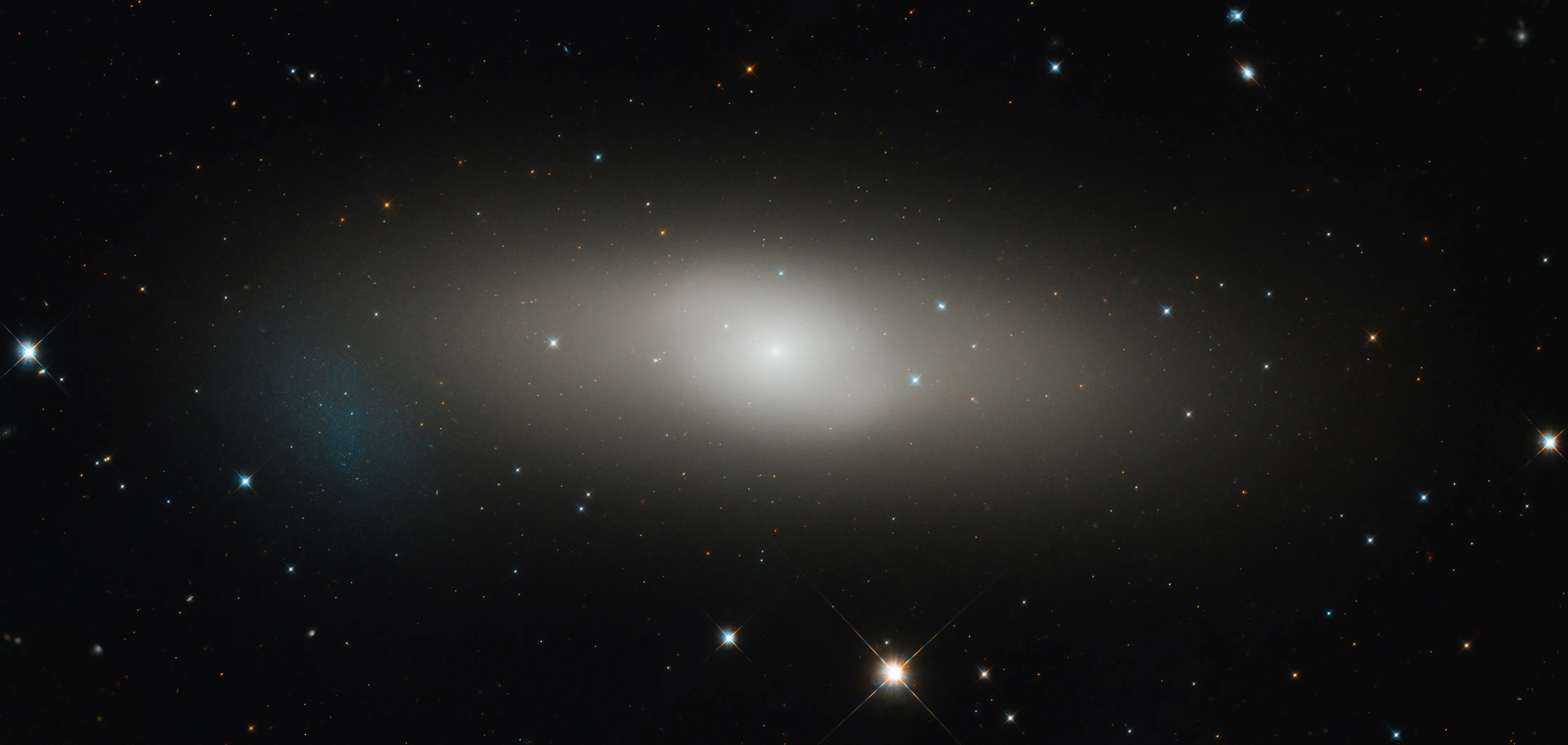 Large lens-like diffuse galaxy with bright core. a cloud of blue gas and stars on its lower left edge