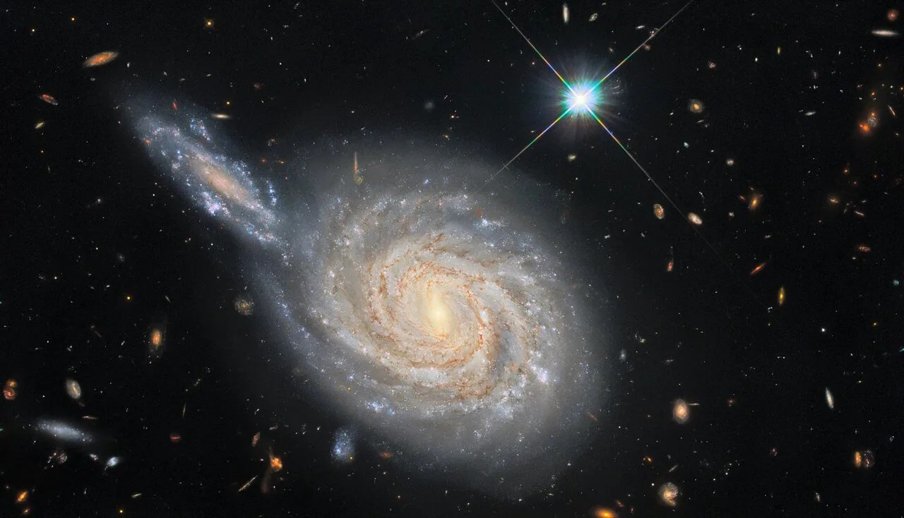 Spiral galaxy at center, with an edge-on galaxy at upper left (10 o'clock) that appears to be crashing into the spiral. bright-blue star to the upper right. many fainter galaxies in the background