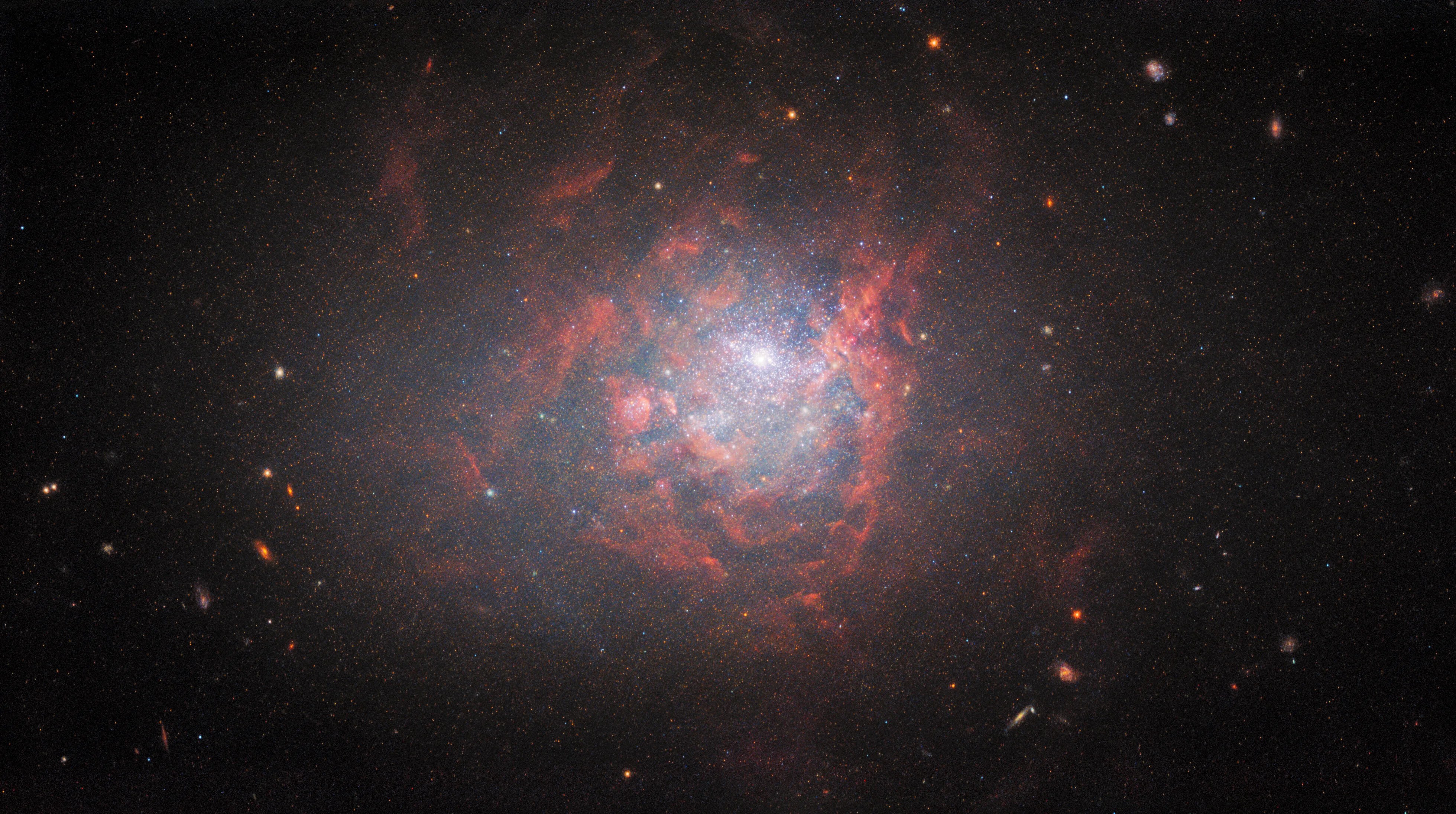 Bright-white star-filled center of the image/galaxy surrounded by billowing reddish clouds, distant galaxies are visible along the periphery of the galaxy