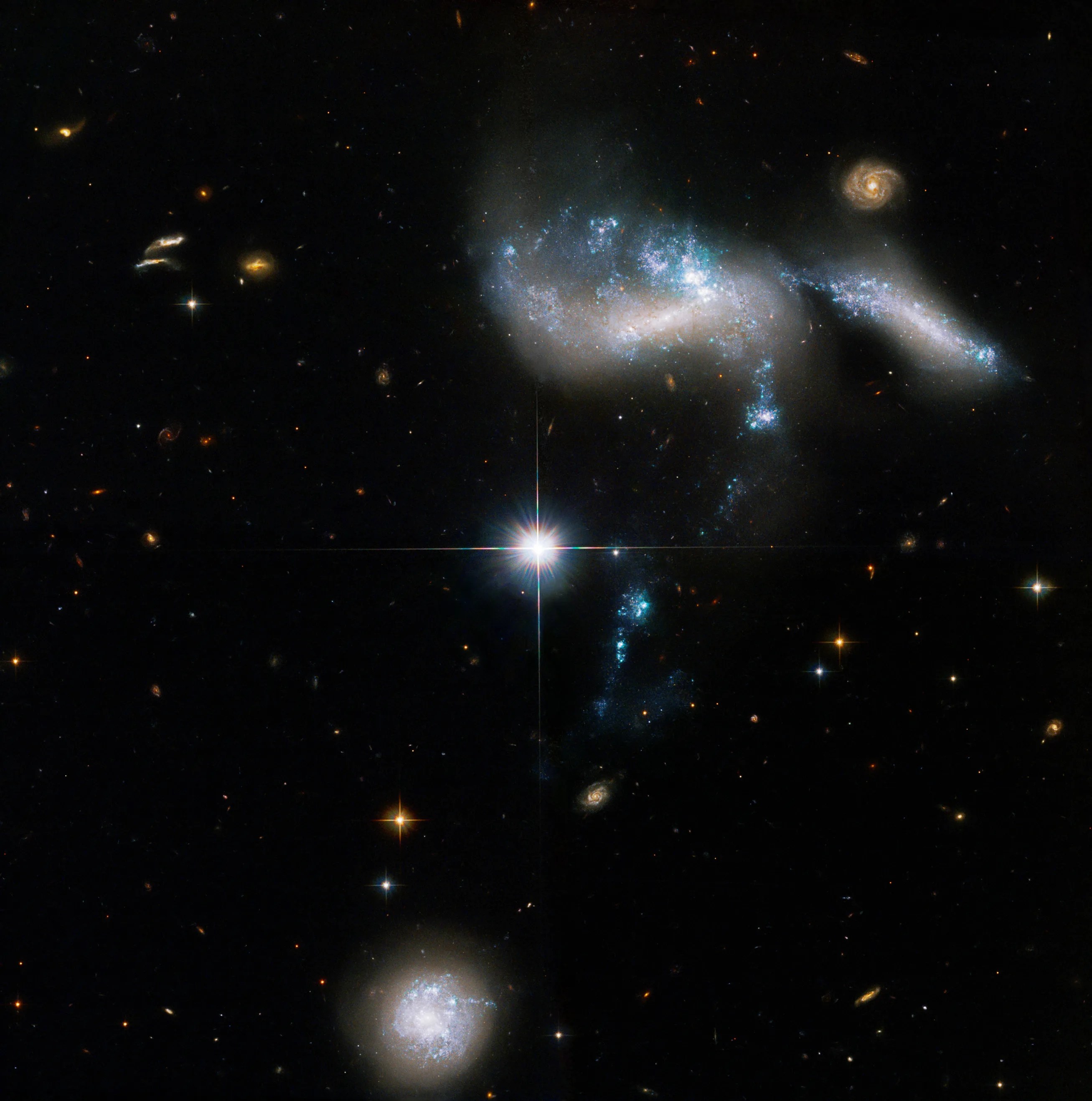 A bright star at image center, above it are two galaxies merging with bright blue star forming regions. a stream of blue-white stars trails off to the bottom of the image just left of center to another dwarf galaxy at the bottom of the image.