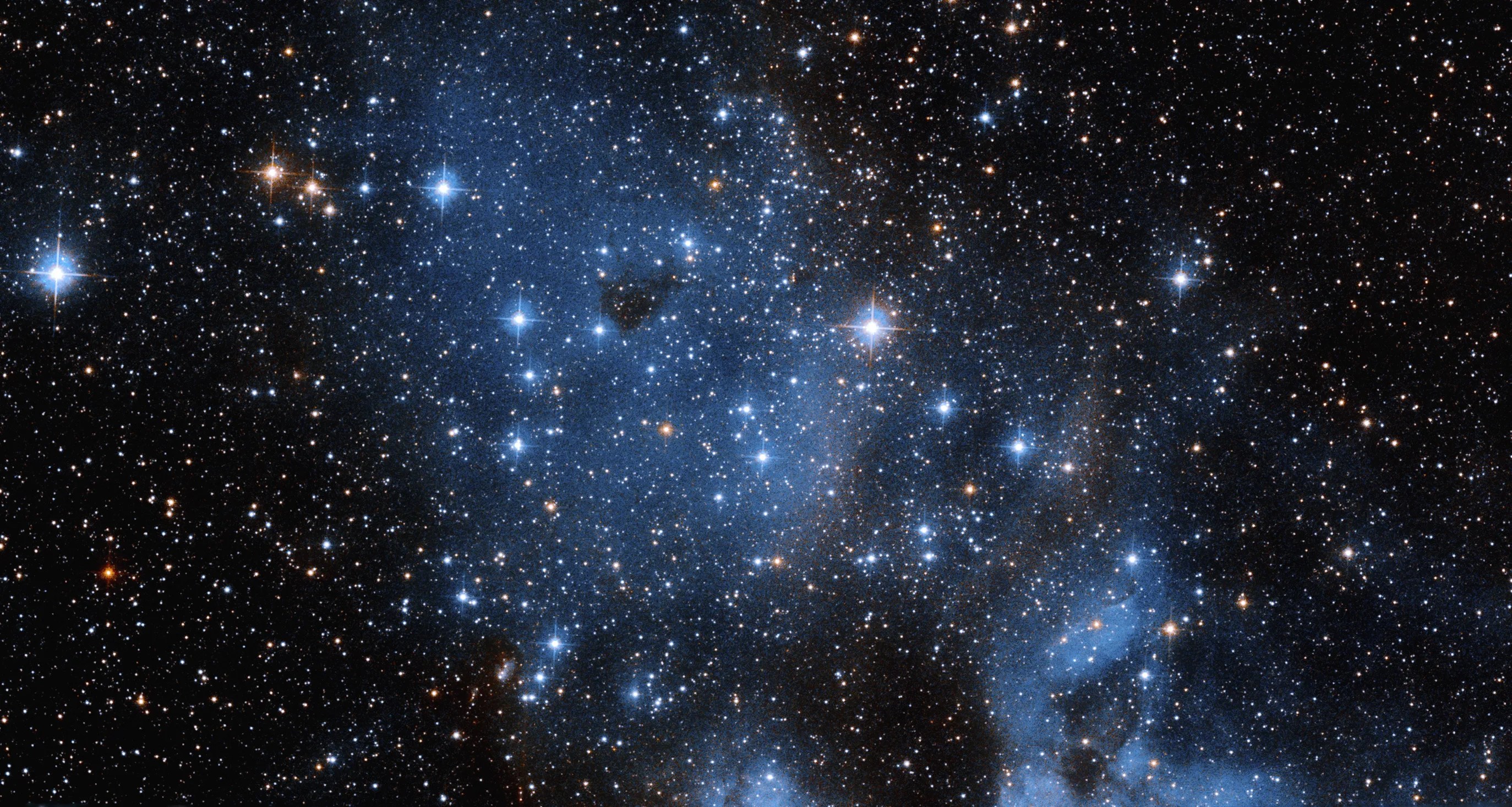 Bright, blue-white stars of varying sizes fil the scene. a smattering of orange-white stars dotted throughout. a swath of pale-blue gas and dust extends from upper left to lower right across the image.