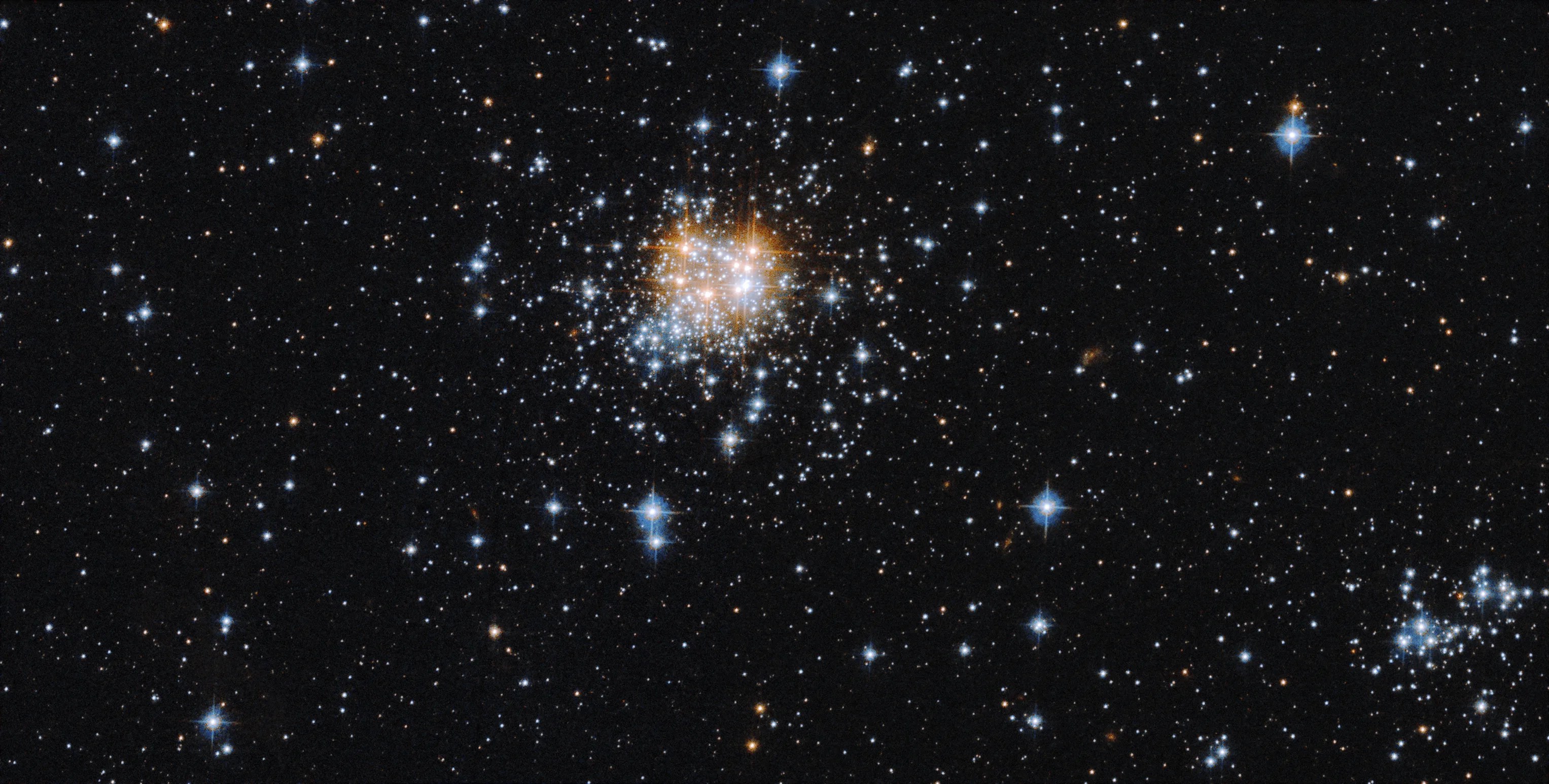 A tight group of bright, blue-white stars just left of center in the image. the group also holds a few orang-white stars. a smattering of blue-white stars against a black background fills the rest of the scene.
