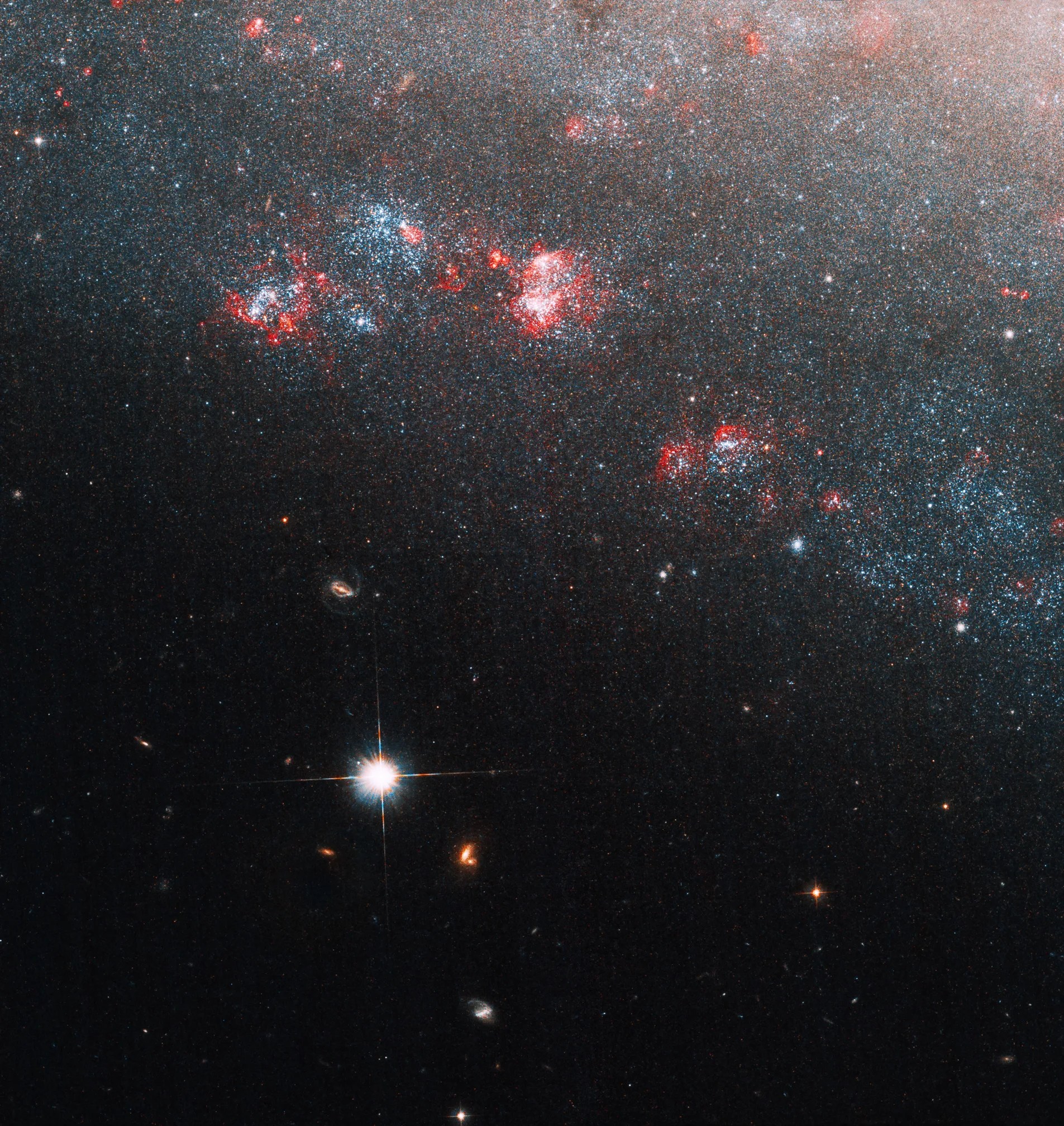 A portion of the galaxy fills the upper right half of the image, blue and red clusters of stars and gas dot its edge, a bright foreground star sits to the lower left of the galaxy