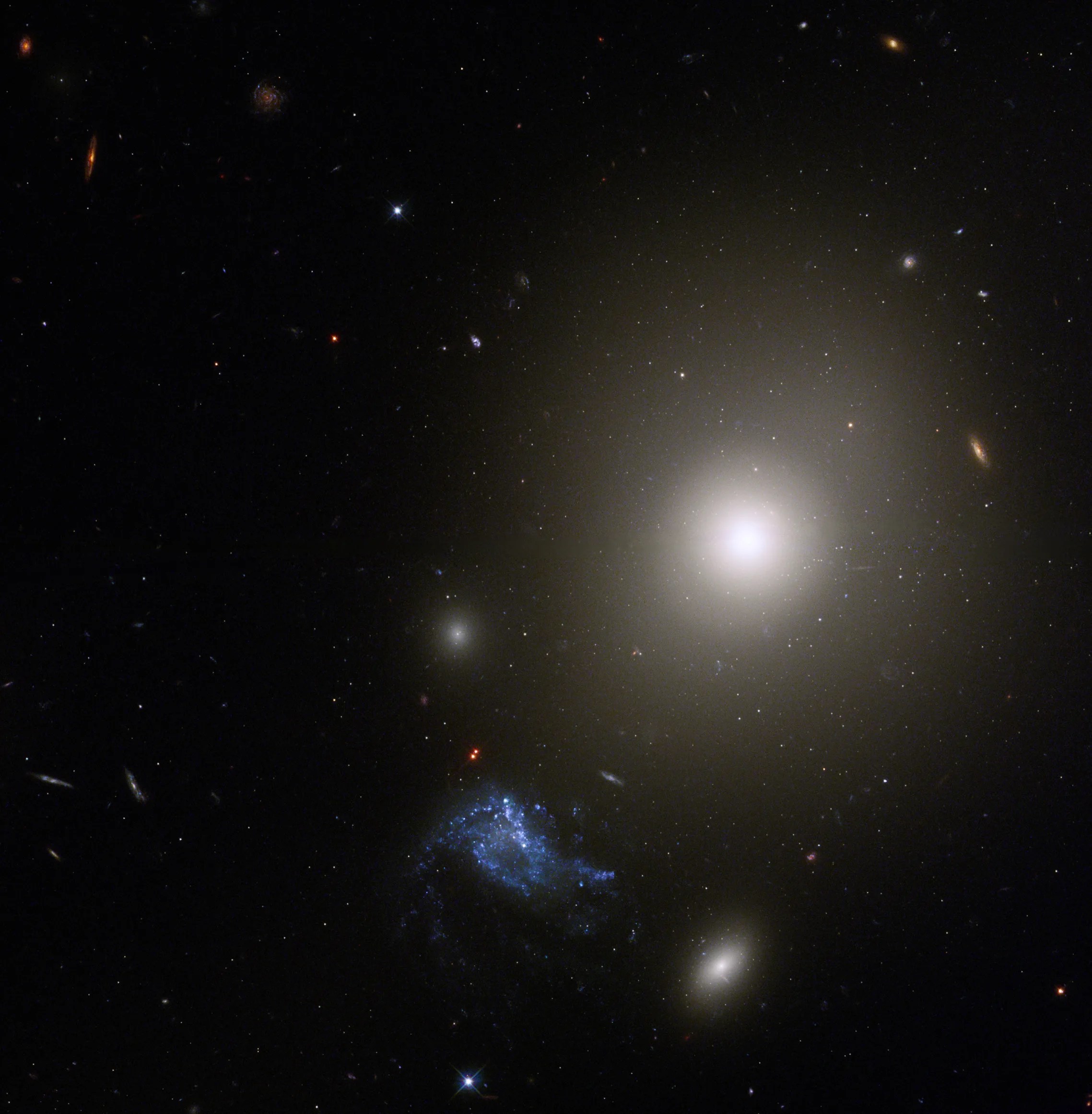 Center, right: bright, white elliptical galaxy with very bright core from which a diffuse glow extends outward. another bright, smaller/more distant galaxy is below it, while bright blue/white irregular is to its lower left.